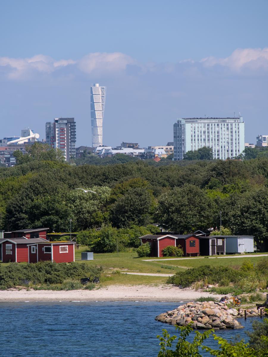 The sea can be seen in the foreground and a few red Swedish cottages on the beach behind it. In the background you can see the Malmö skyline.