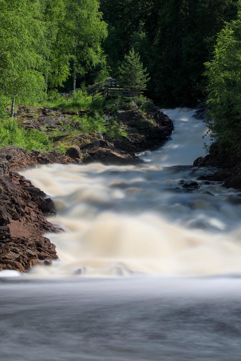 You can see a river with a waterfall. The waterfall is smooth because of long exposure. There is a forest and the blue sky. In the foreground are some rocks.