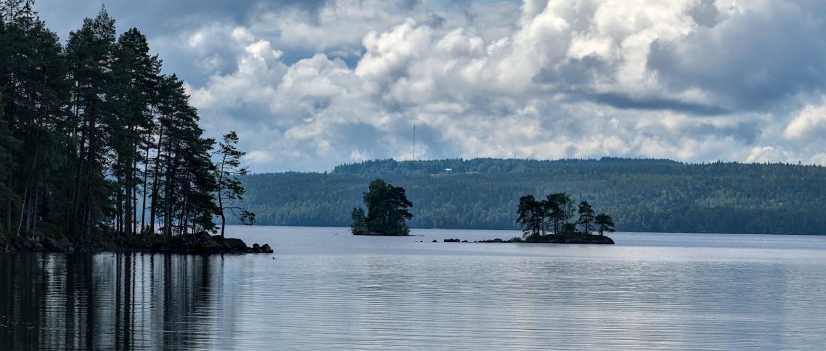 A landscape image of a lake with some small islands on it. In the background there is a forest.