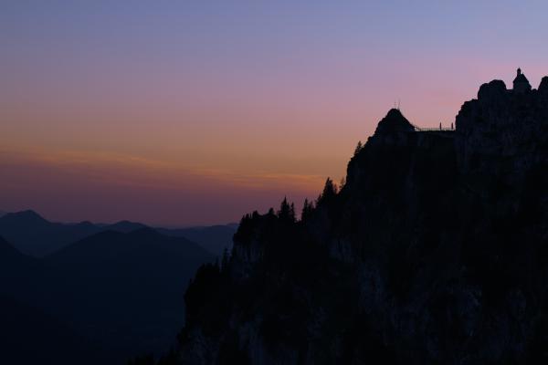 The rugged cliffs of the Wendelstein in the Bavarian Alps stand in dark silhouette against an evening sky fading from violet to pink and orange. Conifers line the ridge, the small Wendelsteinkircherl chapel with its bell turret and cross sits on top, and a railed walkway connects the cliff to a smaller rocky knoll. On the left, layers of blue mountain ridges fade into the haze.
