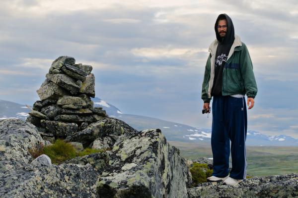 A bearded hiker in glasses, a green jacket, a black hoodie with the hood pulled up and blue track pants stands on lichen-covered rocks beside a summit cairn, holding a compact camera. Beyond lies the Swedish fjäll under an overcast sky, with patches of snow on the distant mountains.
