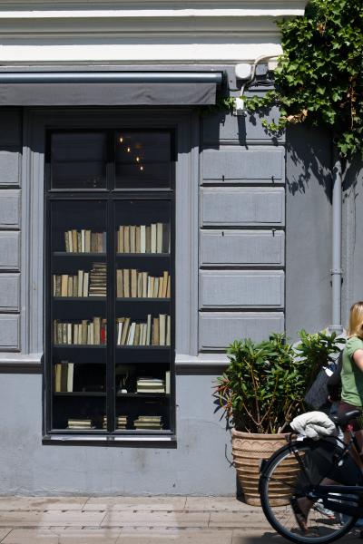 A woman with a bicycle walks past a blue-grey facade with a large window full of old books.