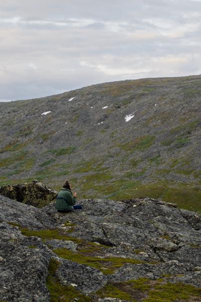 A hiker in a jacket and track pants, his hair in a top knot, smokes in the barren fjäll landscape. Behind him rises a stony mountainside with a few patches of snow. The sky is heavily overcast. The whole scene has a pensive, slightly depressing mood.