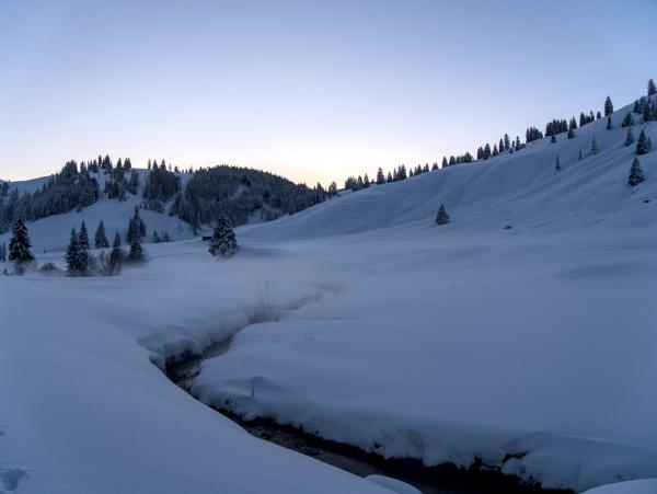 A snow-covered valley in the mountains. There is a small stream in the snow. In the background there are some snow-covered hills.