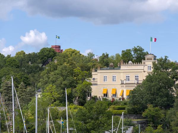 A Swedish flag flying on a castle tower which is hidden behind some trees. More visible is a building with the Italian flag on it.
