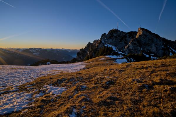 Zu sehen ist der weite Gipfel des Wendelsteins. Im Vordergrund liegt eine teilweise schneebedeckte Wiese. Und im Hintergrund sind einige Berge. Die Sonne geht unter, aber von der linken Seite fallen einige Sonnenstrahlen ins Bild. Der Himmel ist blau und orange.