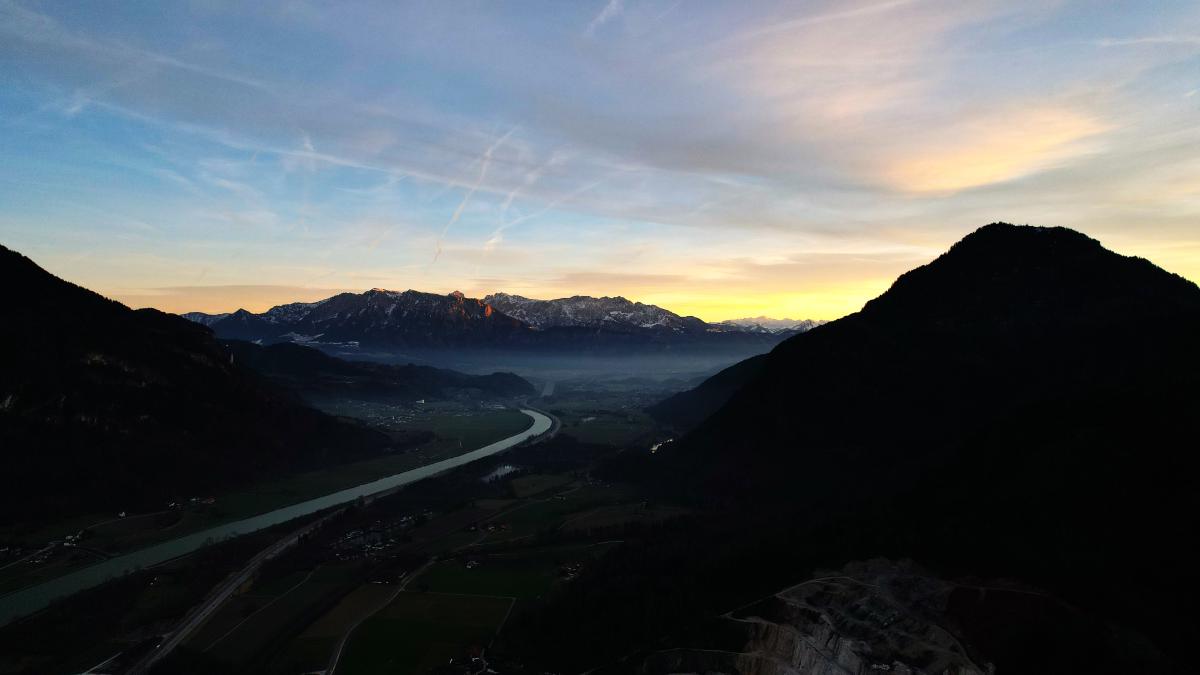 Ein friedlicher Sonnenuntergang über einem sich windenden Fluss in einem österreichischen Tal, mit Bergen im Hintergrund.