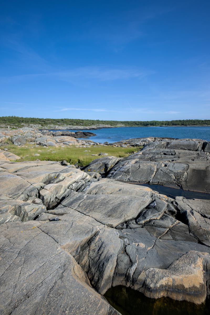 Da ist ein felsiger Strand mit einigen kleinen Tümpeln darin. Zwischen den Felsen ist eine grüne Wiese zu sehen. Dahinter liegt das Meer und dahinter eine Landmasse.