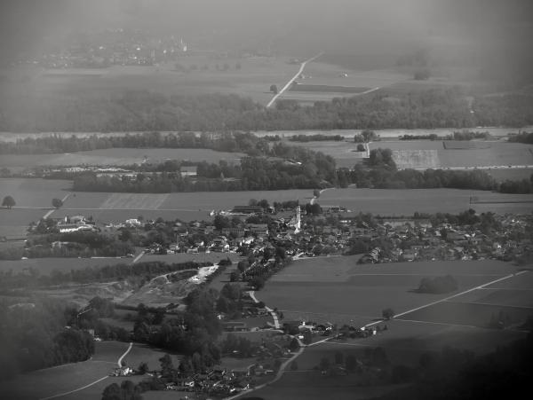 Ein Schwarz-Weiß-Bild eines kleinen Dorfes in Bayern, Deutschland. Das Bild wurde von einem Berg aus durch den Nebel aufgenommen. Hinter dem Dorf sind im Hintergrund ein Fluss und ein weiteres Dorf zu sehen.