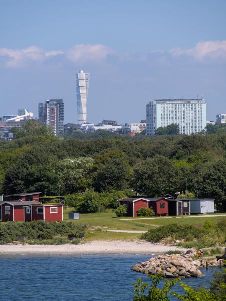 Im Vordergrund ist das Meer zu sehen und dahinter einige rote schwedische Hütten am Strand. Im Hintergrund erkennt man die Skyline von Malmö.