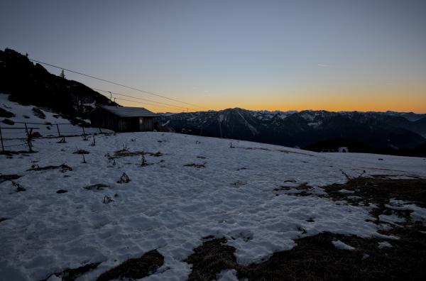 Eine Hütte in den bayerischen Bergen mit einer Wiese davor. Auf der Wiese liegt Schnee, und im Hintergrund erhebt sich eine Bergkette mit einem Sonnenuntergang dahinter.