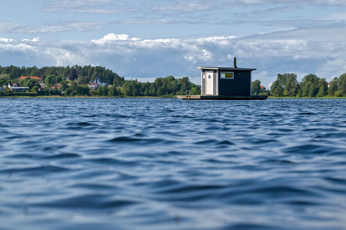 Eine schwimmende Sauna auf einem schwedischen See. Im Hintergrund stehen einige Bäume und Häuser.
