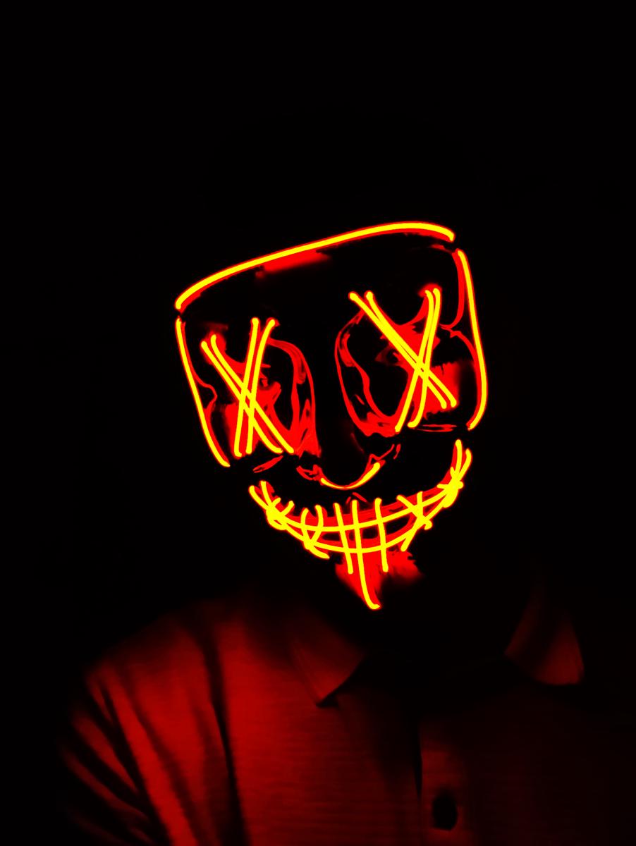 A person in the dark wearing a light-up mask whose glowing red and orange wires form angular crossed eyes and a stitched grin. Only the mask and the collar of a polo shirt are lit.