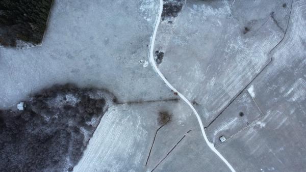 En lodrät flygvy över ett snötäckt landskap: en ljus väg svänger mellan vita fält, en mörk remsa kala träd löper längs vänsterkanten och några små byggnader står i utkanterna.