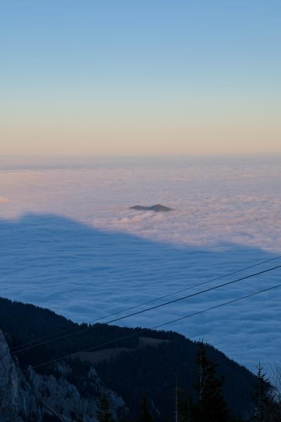Dieses Bild wurde von oberhalb der Wolken aufgenommen. Man blickt von einem Berg auf sie hinab. Im Bild ist ein weiterer Berg zu sehen. Der Himmel ist hellblau / orange.