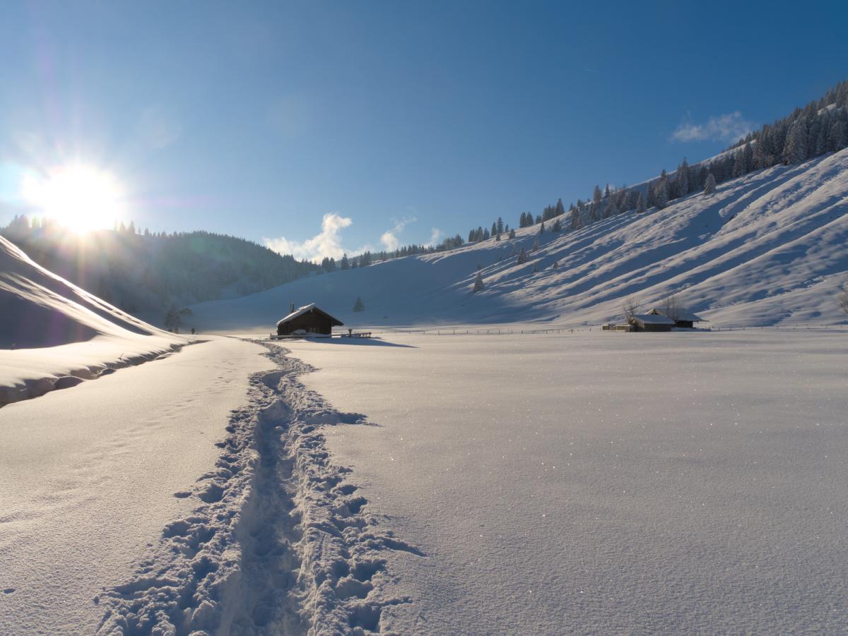 Eine Winterszene in den bayerischen Alpen. Das Bild wurde gegen die Sonne aufgenommen; in einem kleinen Tal zwischen den Bergen ist eine kleine Hütte zu sehen. Es liegt tiefer Schnee, und ein Pfad führt hindurch.