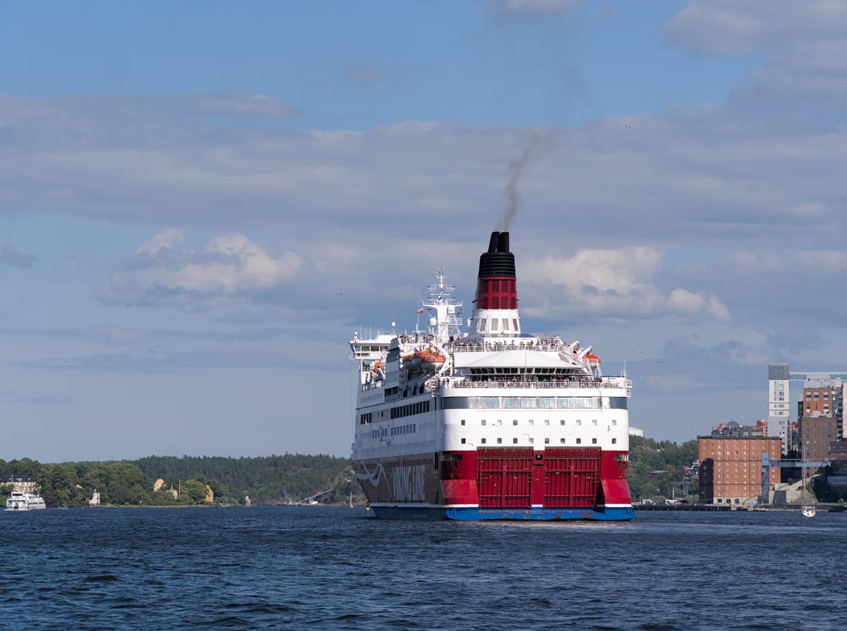 Eine Art Fährschiff, das von Stockholm ablegt. Im Hintergrund sind einige Inseln des Schärengartens zu sehen. Der Himmel ist blau mit wenigen Wolken.