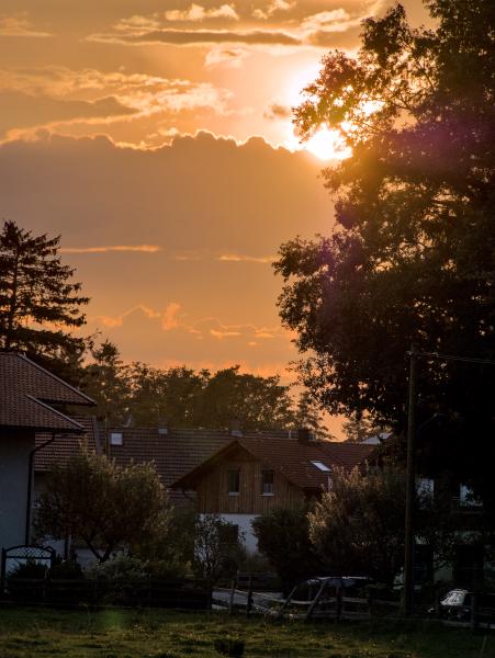 Ein kleines Dorf im Sonnenuntergang. Der bewölkte Himmel ist orange, und die Sonne scheint durch die Bäume.