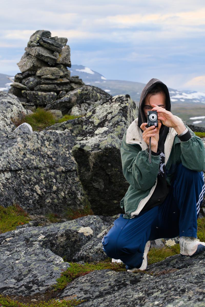 I förgrunden sitter en man med uppdragen luva på huk bland lavklädda stenar och riktar en liten kompaktkamera mot betraktaren. Bakom honom markerar ett röse toppen, och i bakgrunden breder fjällens vida landskap ut sig, med snöfläckar på bergen längre bort.
