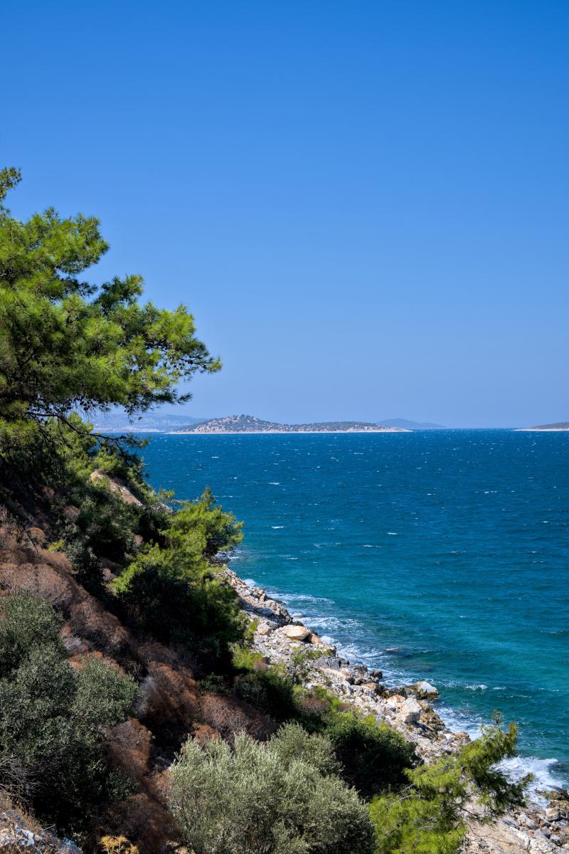 Eine hohe Klippe mit einem steinigen Strand darunter. Das Meer ist türkisblau, und im Hintergrund liegen einige Inseln.