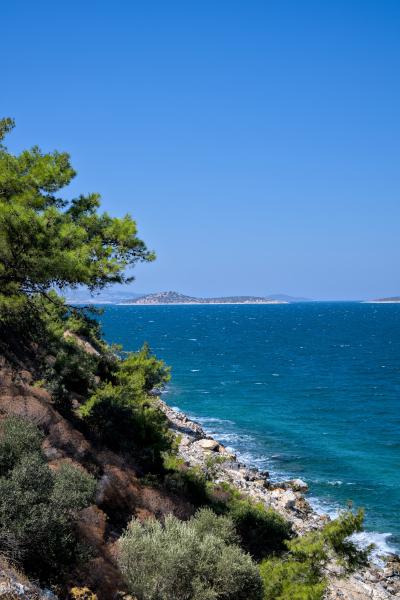 Eine hohe Klippe mit einem steinigen Strand darunter. Das Meer ist türkisblau, und im Hintergrund liegen einige Inseln.
