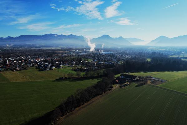 En flygvy över gröna och plöjda fält mot en by; ett pappersbruk med höga skorstenar skickar upp en vit rökpelare, och de disiga alperna kantar horisonten under en blå himmel.