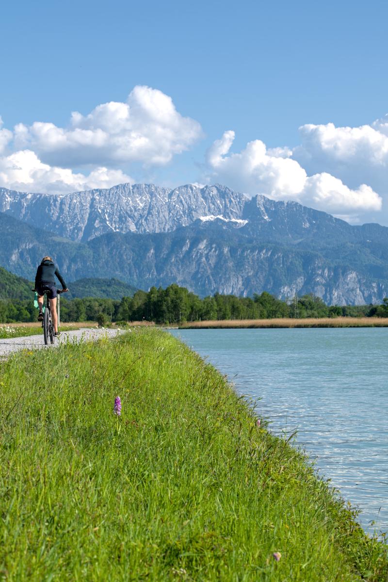 Ein Radfahrer, der an einem Fluss entlangfährt, mit den Bergen im Hintergrund.