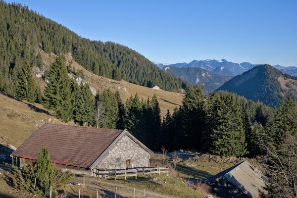 Eine Bergszene mit einer Holzhütte im Vordergrund. Hinter der Hütte liegt ein Wald, und es sind einige weitere Berge zu sehen. Der Himmel ist blau.