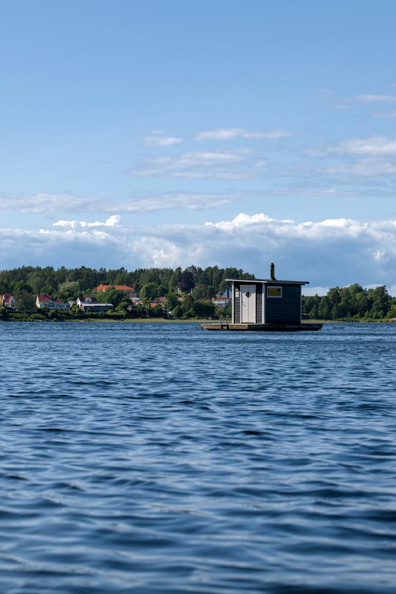 Eine schwimmende Sauna auf einem schwedischen See. Im Hintergrund stehen einige Bäume und Häuser.