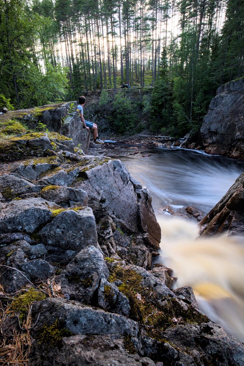 Ein Selbstporträt von mir, wie ich auf einem Felsen neben einem Wasserfall sitze. Der Wasserfall ist wegen der Langzeitbelichtung verwaschen.