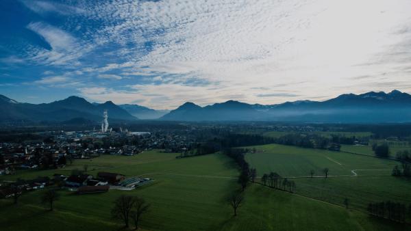 En flygvy över gröna fält och en by med en fabriksskorsten; bakom dem ligger en dimbank vid alpernas fot under en himmel strimmig av moln.