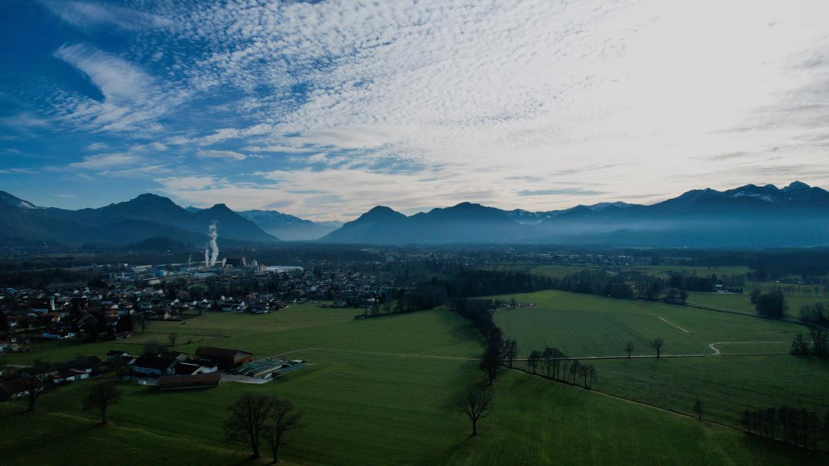 En flygvy över gröna fält och en by med en fabriksskorsten; bakom dem ligger en dimbank vid alpernas fot under en himmel strimmig av moln.