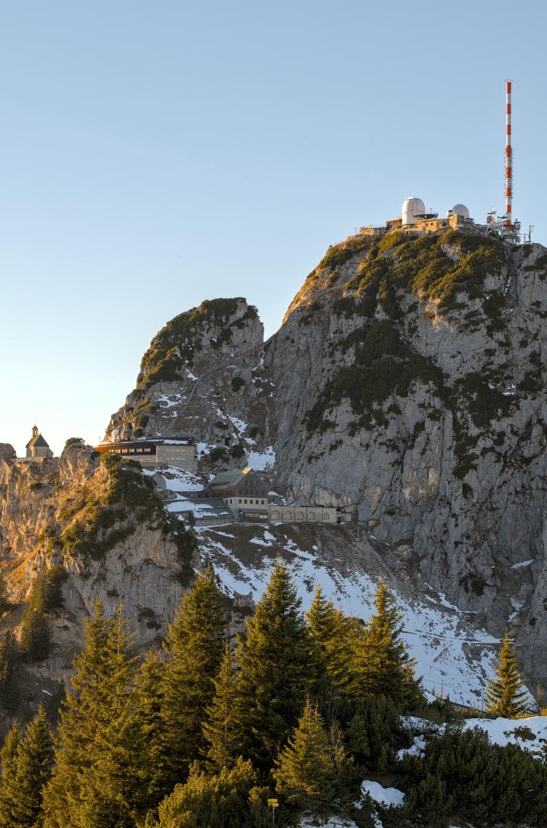 Auf diesem Bild ist der Gipfel eines hohen Berges mit einer Art Antenne darauf zu sehen. Unterhalb des Gipfels stehen einige Gebäude, und am unteren Bildrand liegt ein kleiner Wald.