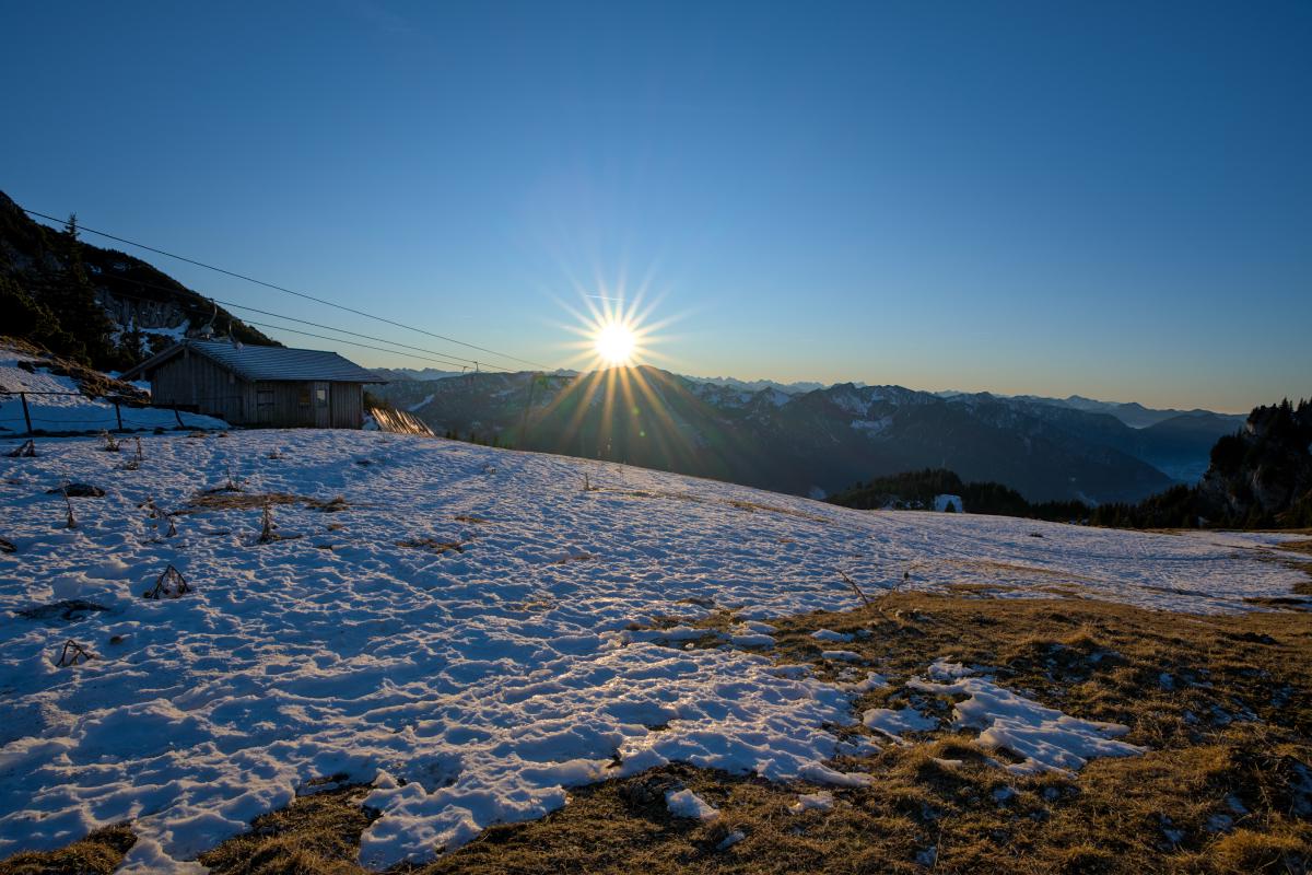 Das Bild zeigt eine Szene in einer Berglandschaft. Die Sonne geht direkt über einigen Bergen unter. Auf der linken Seite stehen eine Berghütte und ein Skilift. Im Vordergrund liegt eine Wiese, auf der stellenweise Schnee liegt.