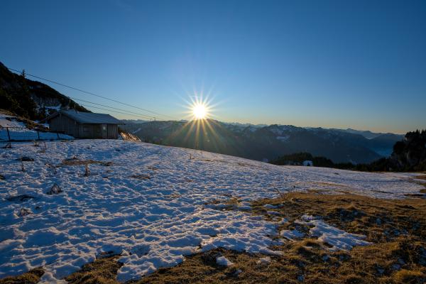 Das Bild zeigt eine Szene in einer Berglandschaft. Die Sonne geht direkt über einigen Bergen unter. Auf der linken Seite stehen eine Berghütte und ein Skilift. Im Vordergrund liegt eine Wiese, auf der stellenweise Schnee liegt.