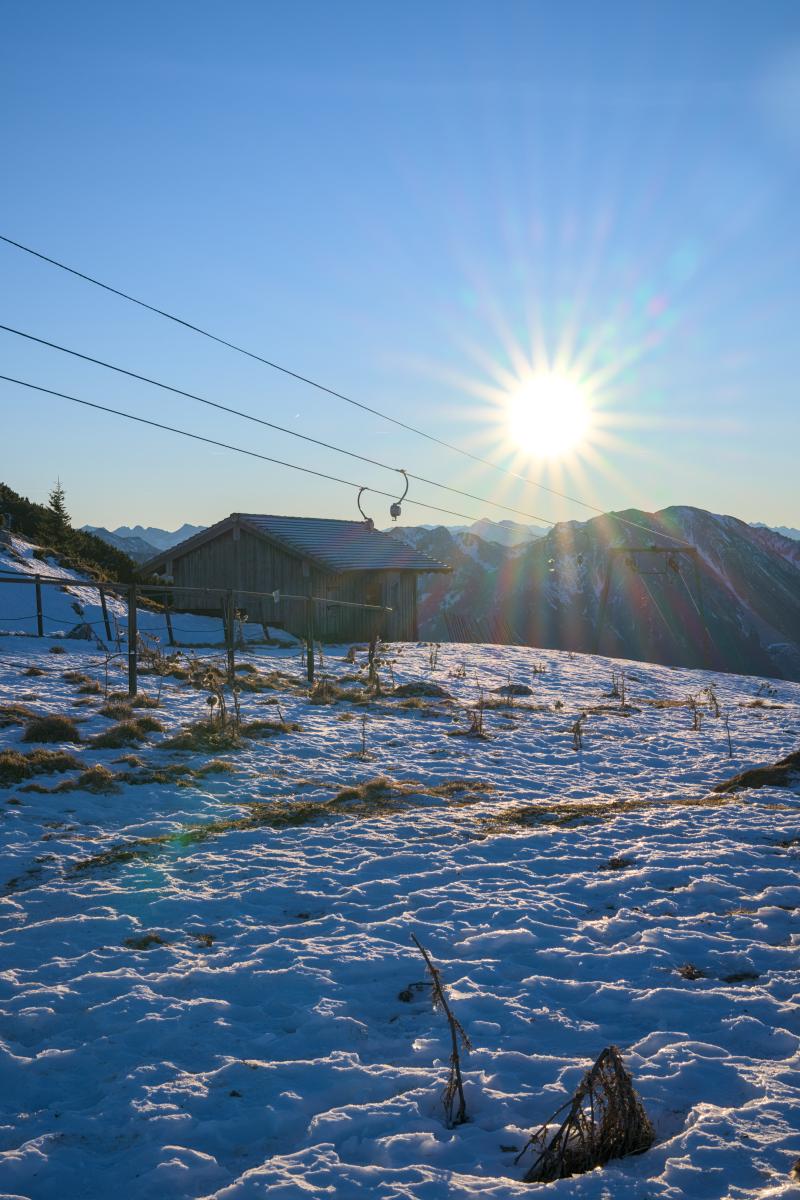 Eine Szene, aufgenommen in den bayerischen Bergen. Im Vordergrund liegt eine sonnige Wiese mit einer Berghütte und einem Skilift darauf. Im Hintergrund ist die Sonne mit einem Sonnenstern direkt über einer Bergkette zu sehen. Der Himmel ist blau und am Horizont ein wenig orange.