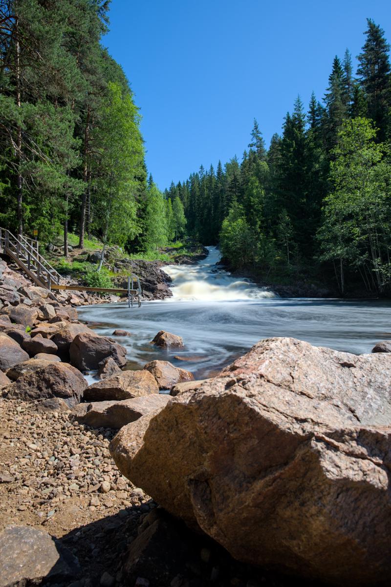 Zu sehen ist ein Fluss mit einem Wasserfall. Der Wasserfall wirkt wegen der Langzeitbelichtung glatt. Dazu ein Wald und der blaue Himmel. Im Vordergrund liegen einige Felsen.