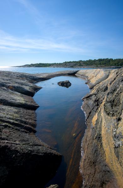 In der Mitte des Bildes ist ein kleiner Tümpel aus Meerwasser zu sehen. In seiner Mitte liegt ein Stein. Im Hintergrund ist Land zu erkennen, und dazwischen liegt das Meer.