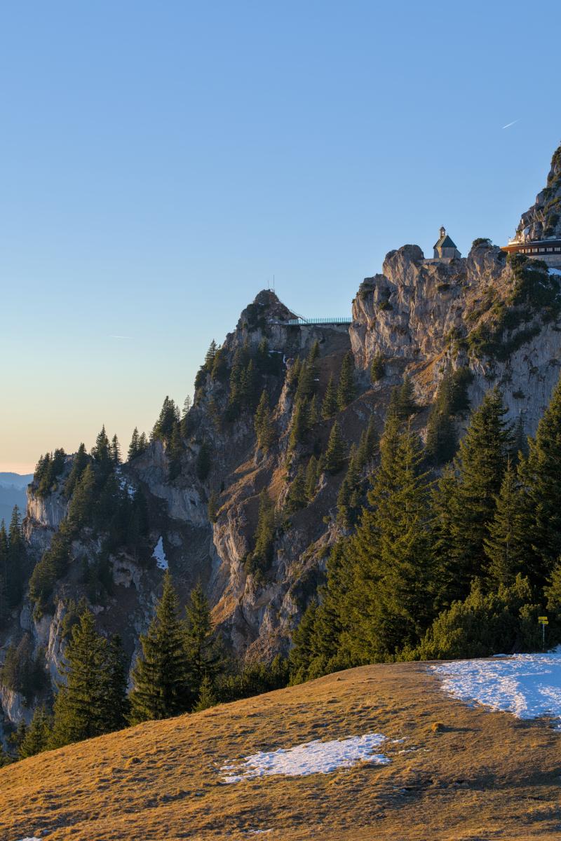 You can see a part of the top of the mountain Wendelstein in the Bavarian Alps. Especially a little church and the rough rocks of the mountain top. In the foreground there is a meadow and a little forest. The sky is blue / orange.
