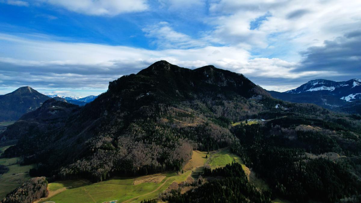 En flygvy över ett skogklätt berg med klippig topp, ängar och stigar i de nedre sluttningarna, och snöpudrade alptoppar under en molnstrimmig blå himmel bakom.