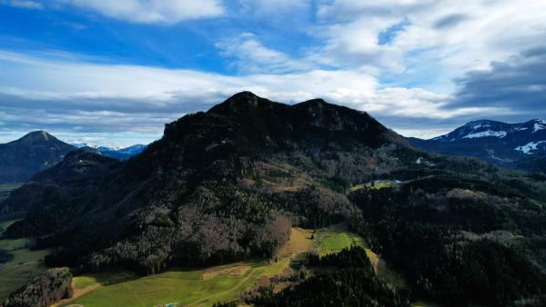 En flygvy över ett skogklätt berg med klippig topp, ängar och stigar i de nedre sluttningarna, och snöpudrade alptoppar under en molnstrimmig blå himmel bakom.