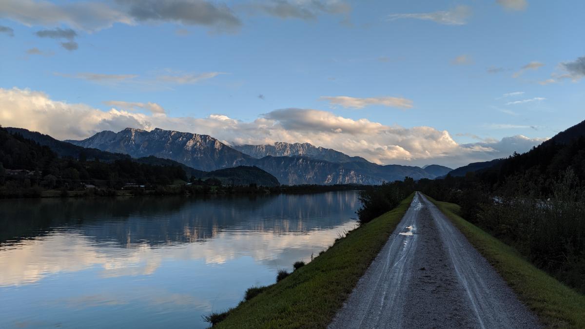 Ein ruhiger Fluss, der den Himmel spiegelt, mit einem Weg daneben und Bergen in der Ferne unter einem bewölkten Himmel.