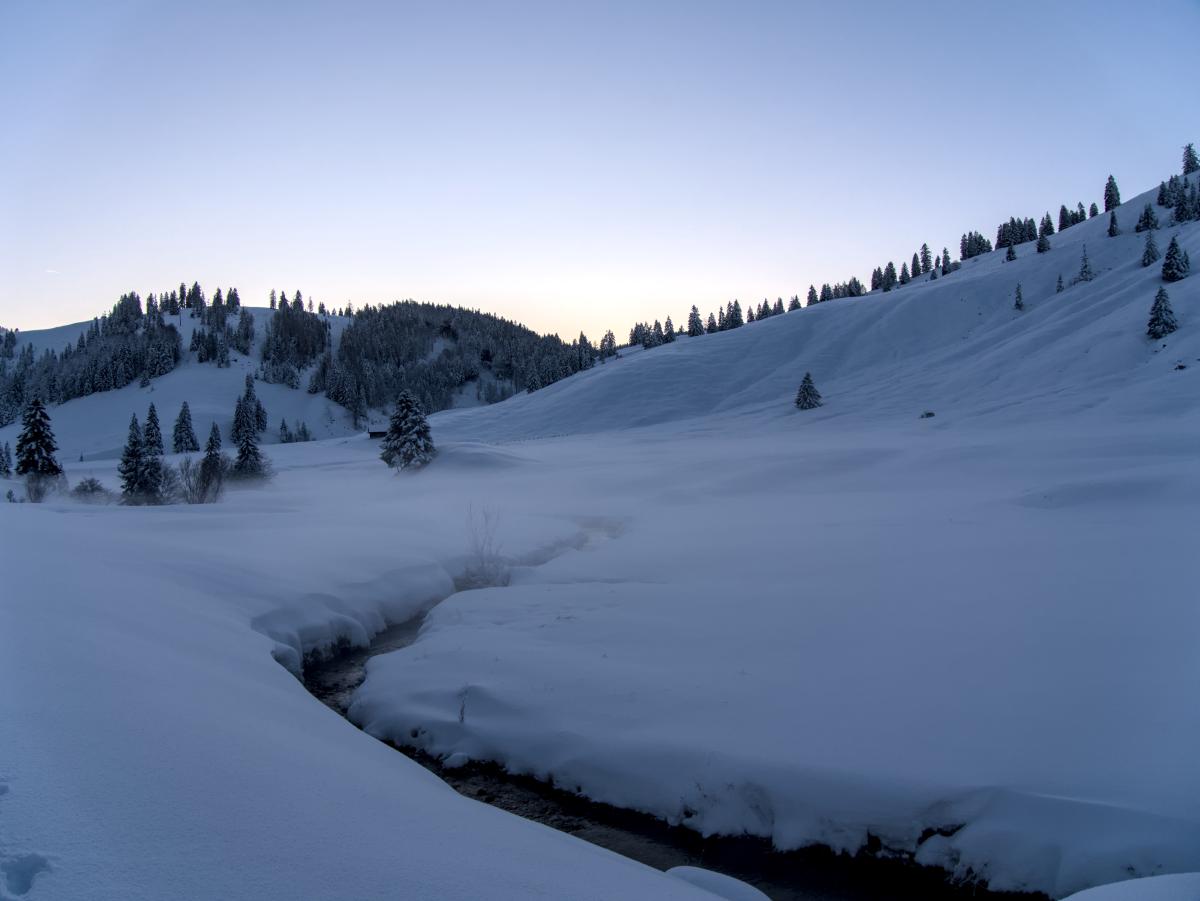 Ein schneebedecktes Tal in den Bergen. Im Schnee verläuft ein kleiner Bach. Im Hintergrund liegen einige schneebedeckte Hügel.