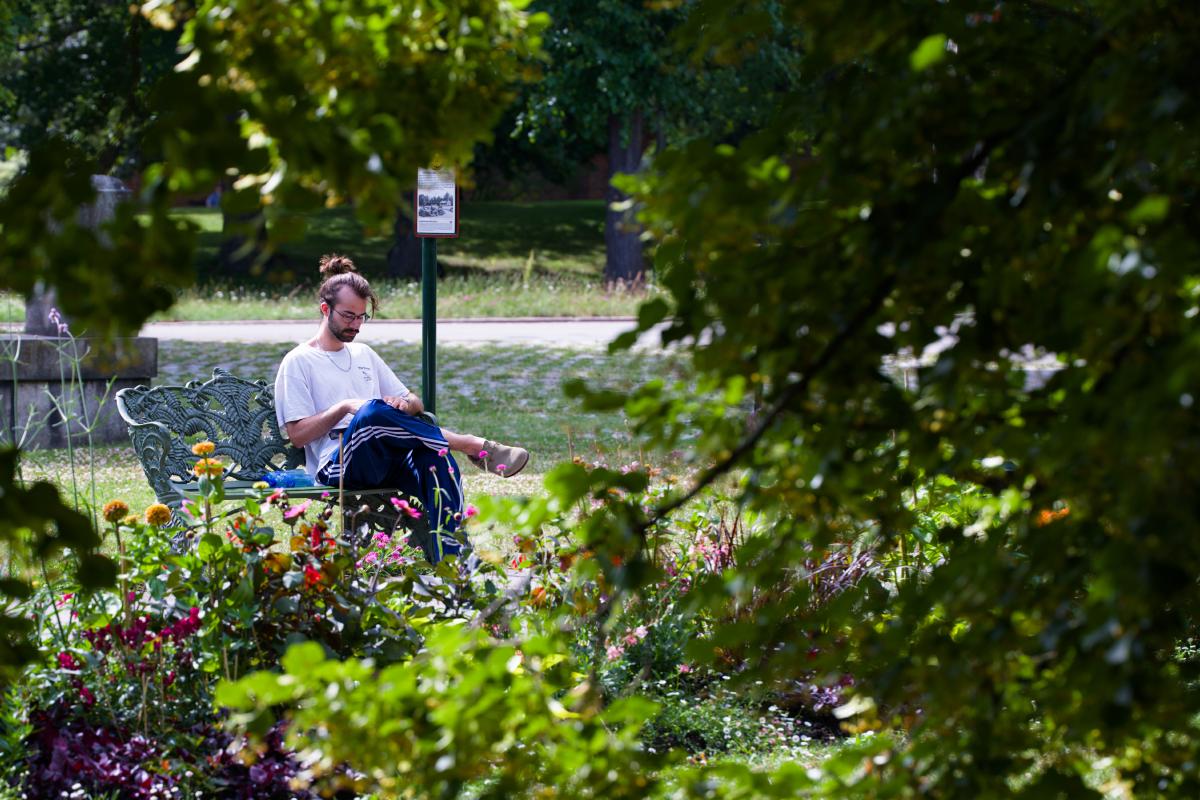 En man i vit t-shirt med glasögon, halskedja och hårknut sitter på en utsmyckad parkbänk av gjutjärn i Stockholm och tittar ner i knät. Han ramas in av ett träds grenar och parkens blommor i förgrunden.