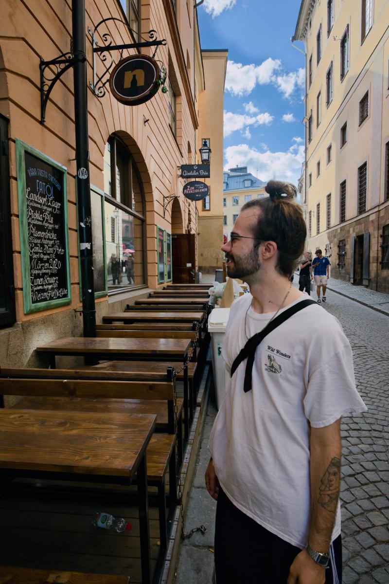 Ein lächelnder Mann mit Bart, Brille und Dutt steht vor einem Pub in Gamla stan, der Altstadt Stockholms, und blickt auf eine Kreidetafel mit schwedischen Fassbieren. Hinter ihm verläuft eine schmale Kopfsteinpflastergasse zwischen gelben und orangefarbenen Häusern unter einem blauen Himmel mit weißen Wolken.