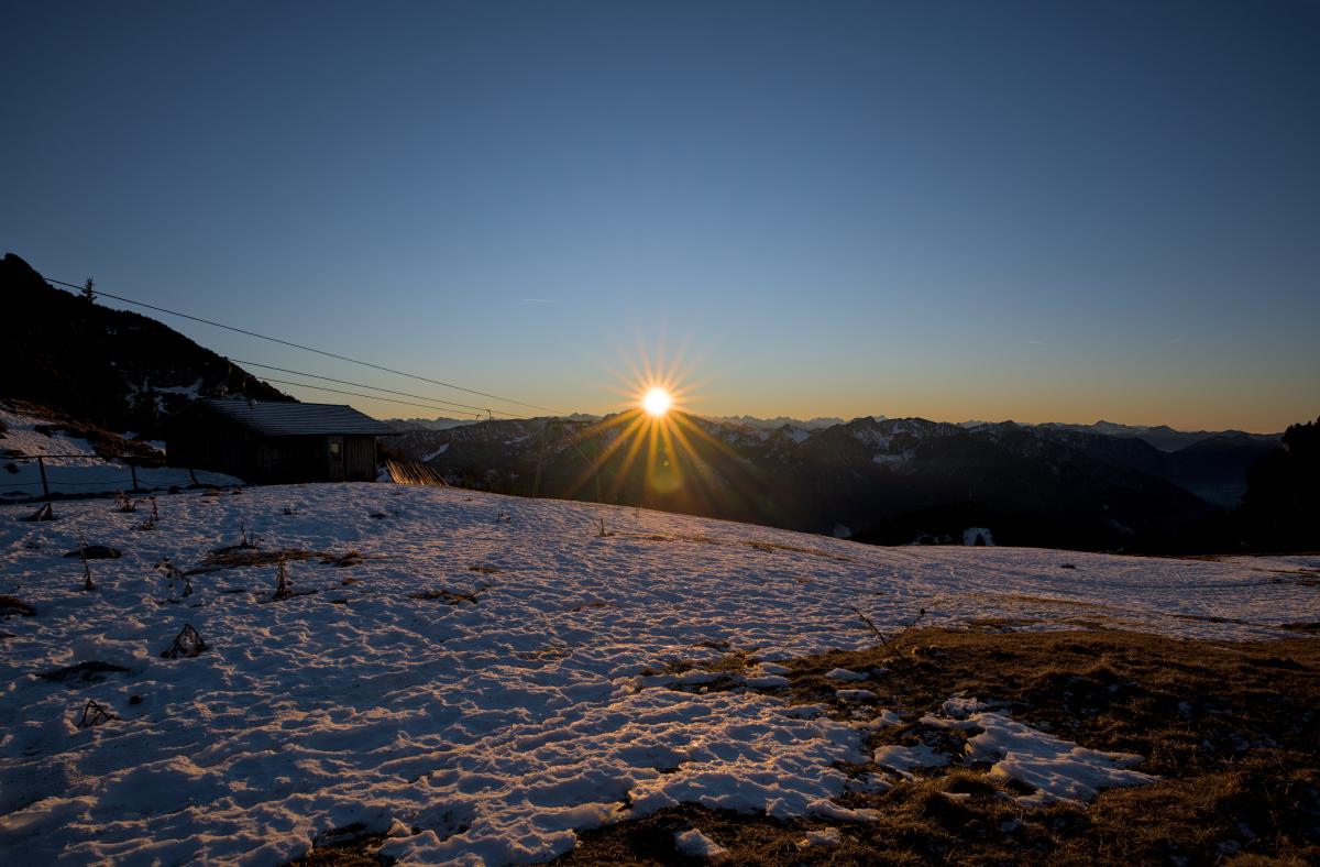 Zu sehen ist der Blick auf eine Bergkette, fotografiert von einem anderen Berg aus. Die Sonne geht in der Richtung unter, in die das Foto aufgenommen wurde. Auf der linken Seite steht eine Berghütte.