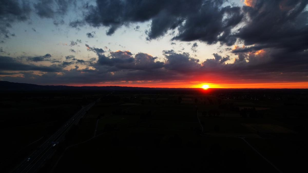 Eine Luftaufnahme bei Sonnenuntergang über flachem Ackerland: eine Autobahn verläuft von links unten in die Ferne, die Sonne steht rot am Horizont und schwere dunkle Wolken füllen die obere Bildhälfte.