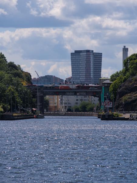 Das Foto wurde von einem Boot aus aufgenommen und zeigt das Meer, eine Brücke im Mittelgrund und einige große Gebäude im Hintergrund. Die Aufnahme entstand in Stockholm.