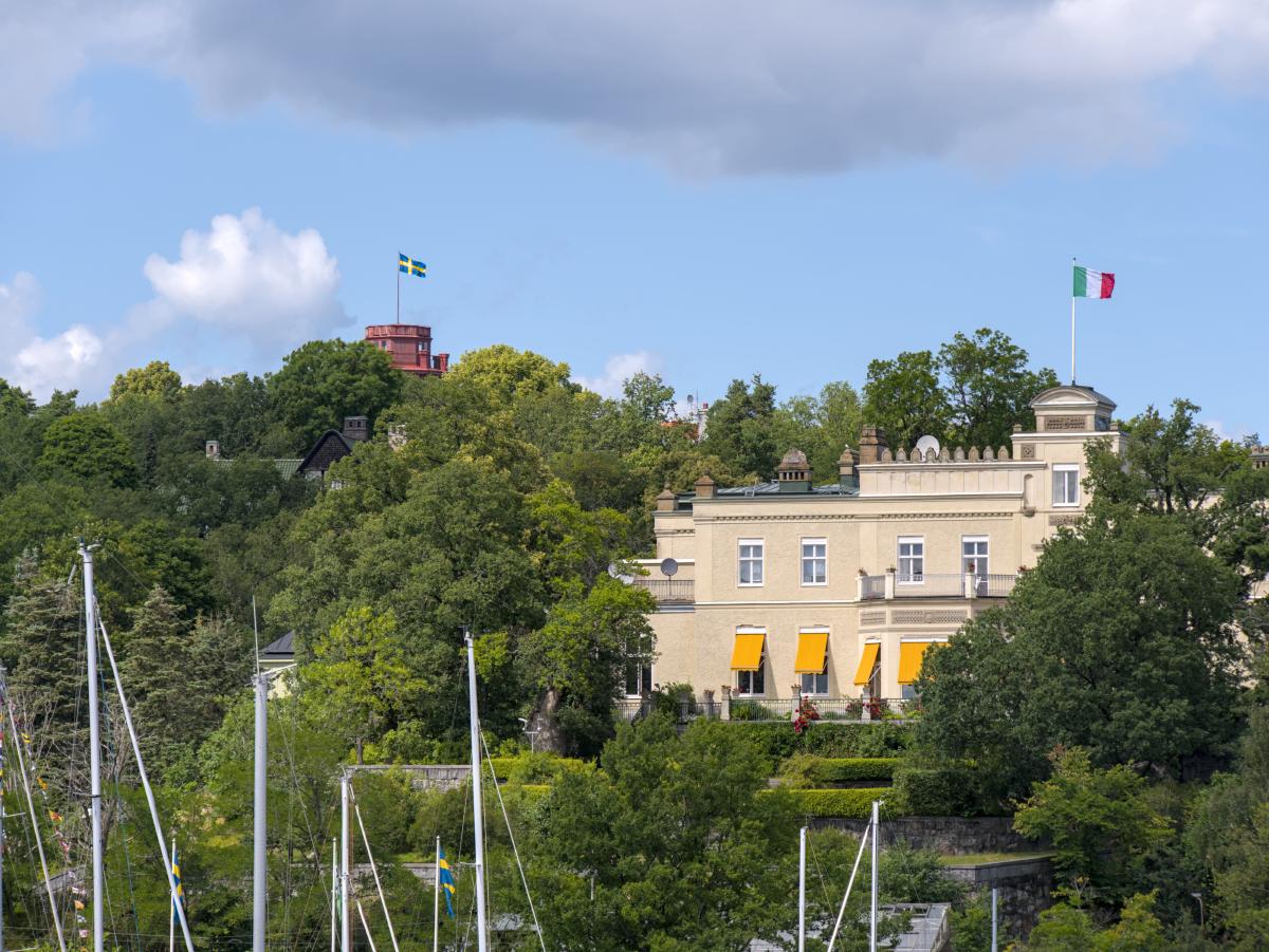 Eine schwedische Flagge, die auf einem Schlossturm weht, der hinter einigen Bäumen verborgen ist. Deutlicher zu sehen ist ein Gebäude mit der italienischen Flagge daran.