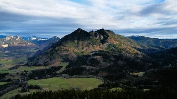 En flygvy över ett mörkt skogklätt berg med dubbeltopp som reser sig över gröna ängar och gårdar, med de snötäckta alperna utsträckta längs horisonten.